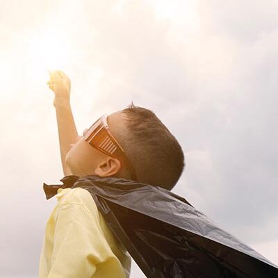 child wearing a cape and sunglasses, hand pointed up to the sun
